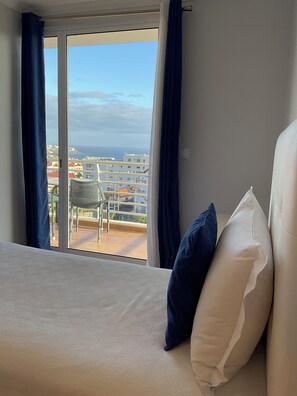 Ett hotellrum med en säng, två kuddar och utsikt över havet och byggnaderna genom ett stort fönster med gardiner.