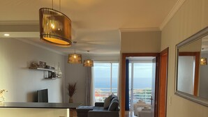 Ett modernt vardagsrum med en soffa, ett matbord och stolar. Genom stora fönster har man utsikt över havet.