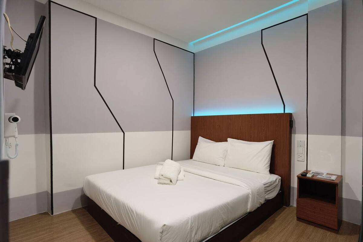 Moderni hotellihuone, jossa on suuri sänky, puinen pääty, yöpöytä lampun kanssa ja seinälle asennettu valvontakamera.