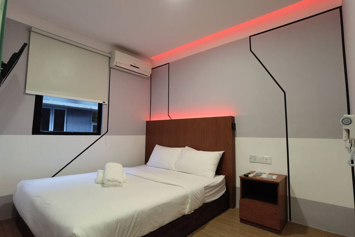 Moderni hotellihuone, jossa on sänky, yöpöytä ja ikkuna, jossa on sälekaihtimet.