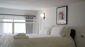 Un lit bien fait, avec du linge de lit blanc et une serviette pliée, un luminaire fixé au mur et un tableau encadré accroché au mur.