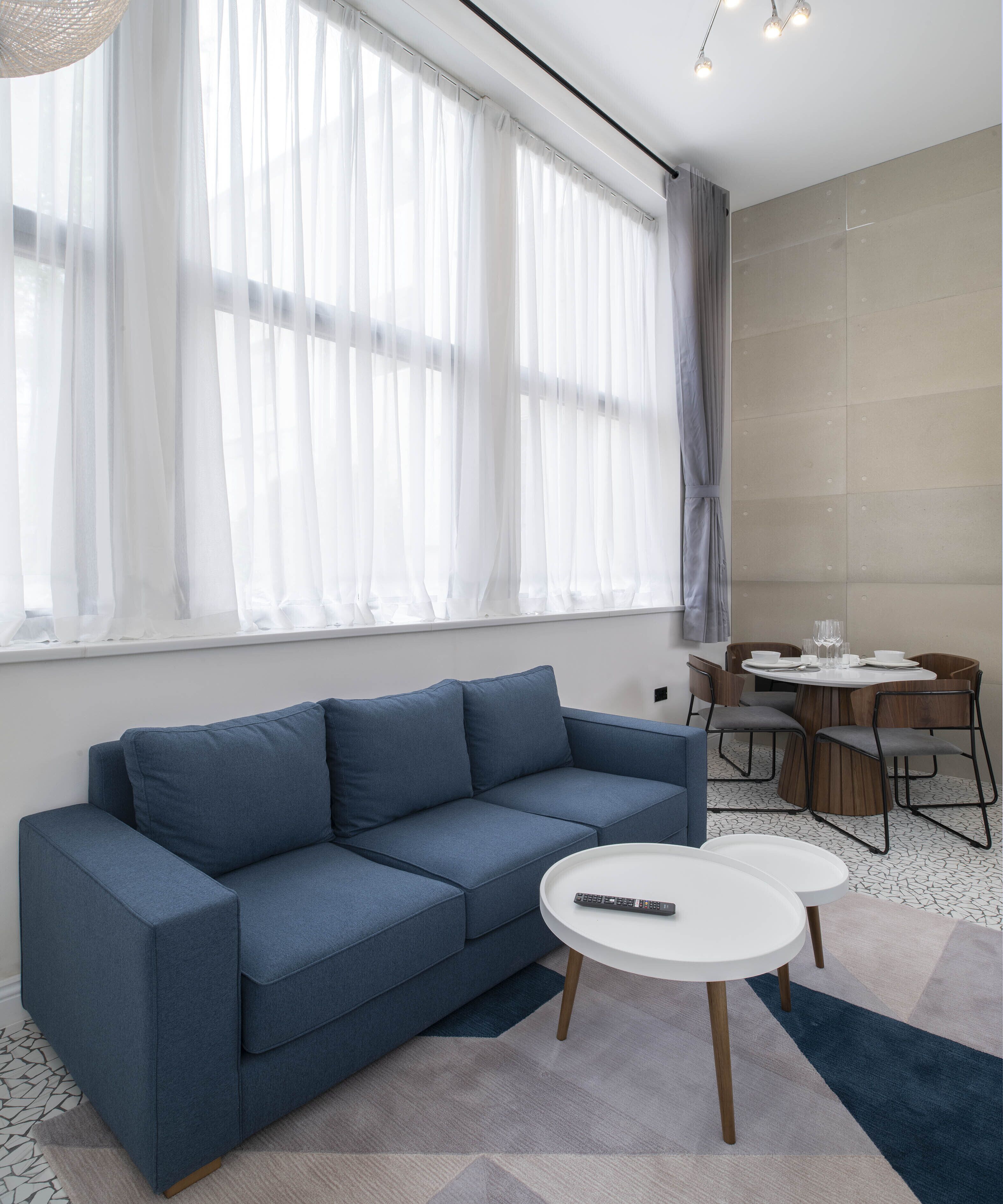 A modern living room with a blue sofa, a dining table with chairs, and a large window with sheer curtains.