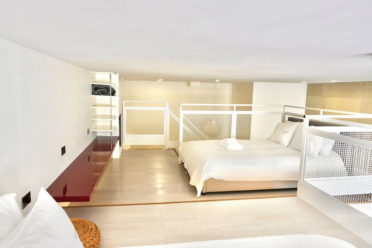 A bedroom with a bed, a small seating area, a staircase, and a view of the outside.