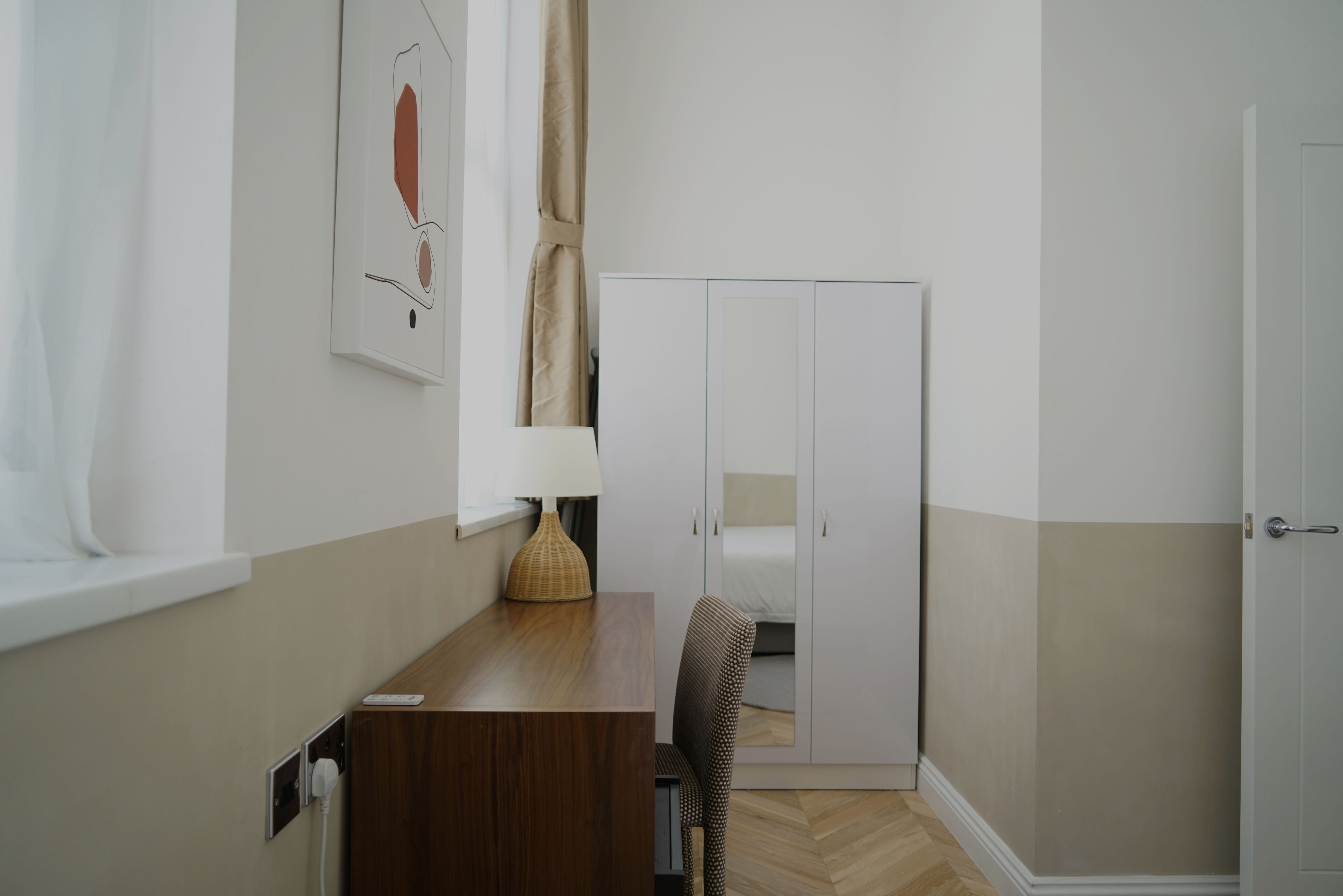 Une chambre d’hôtel de taille modeste, comprenant un bureau en bois, un lit simple, une armoire et une fenêtre avec des rideaux.