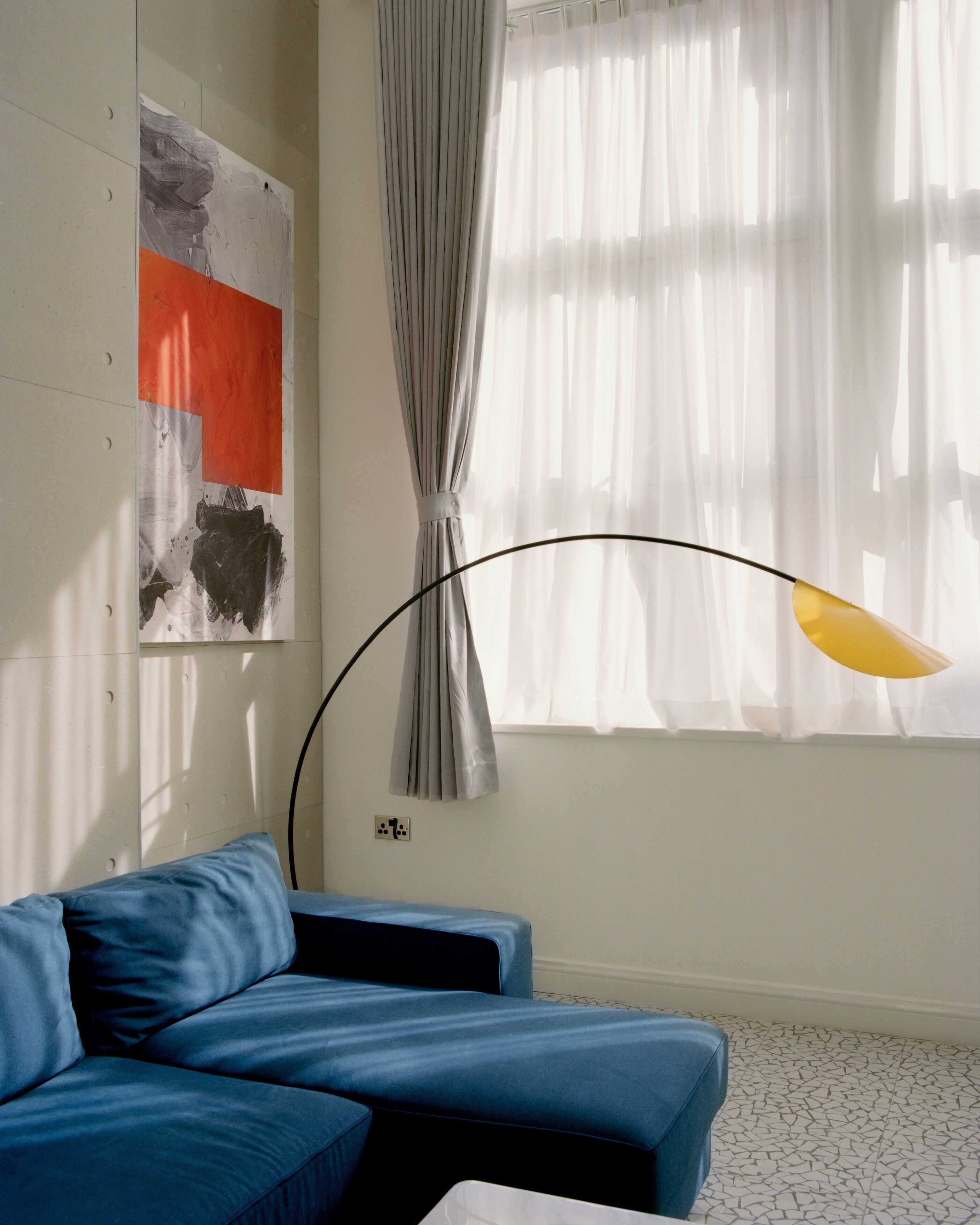 A modern living room with a blue sofa, a painting on the wall, and a window with sheer curtains.
