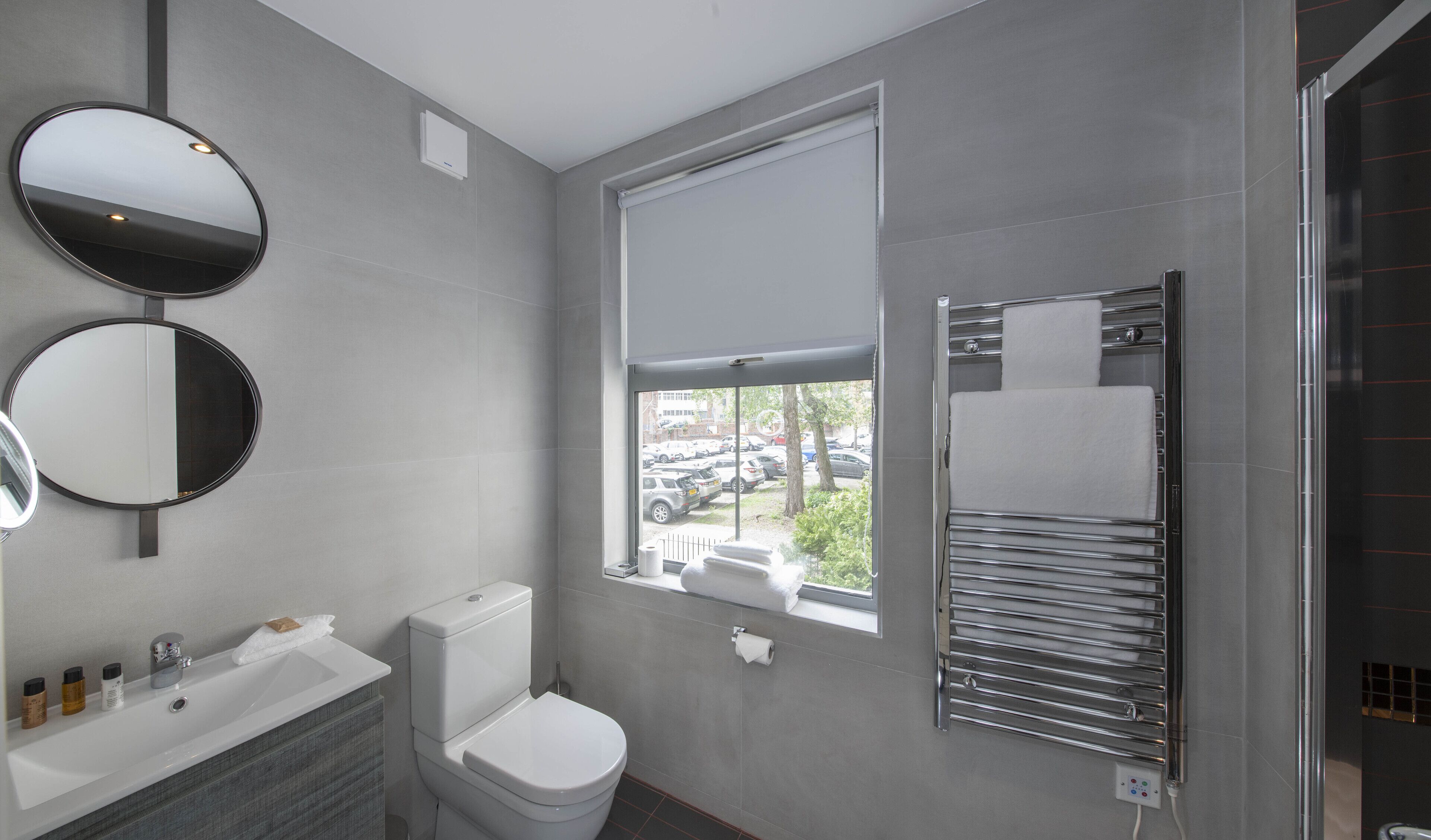 Un baño moderno con inodoro, lavabo y dos espejos. Se ve una ventana con vistas a coches estacionados afuera.
