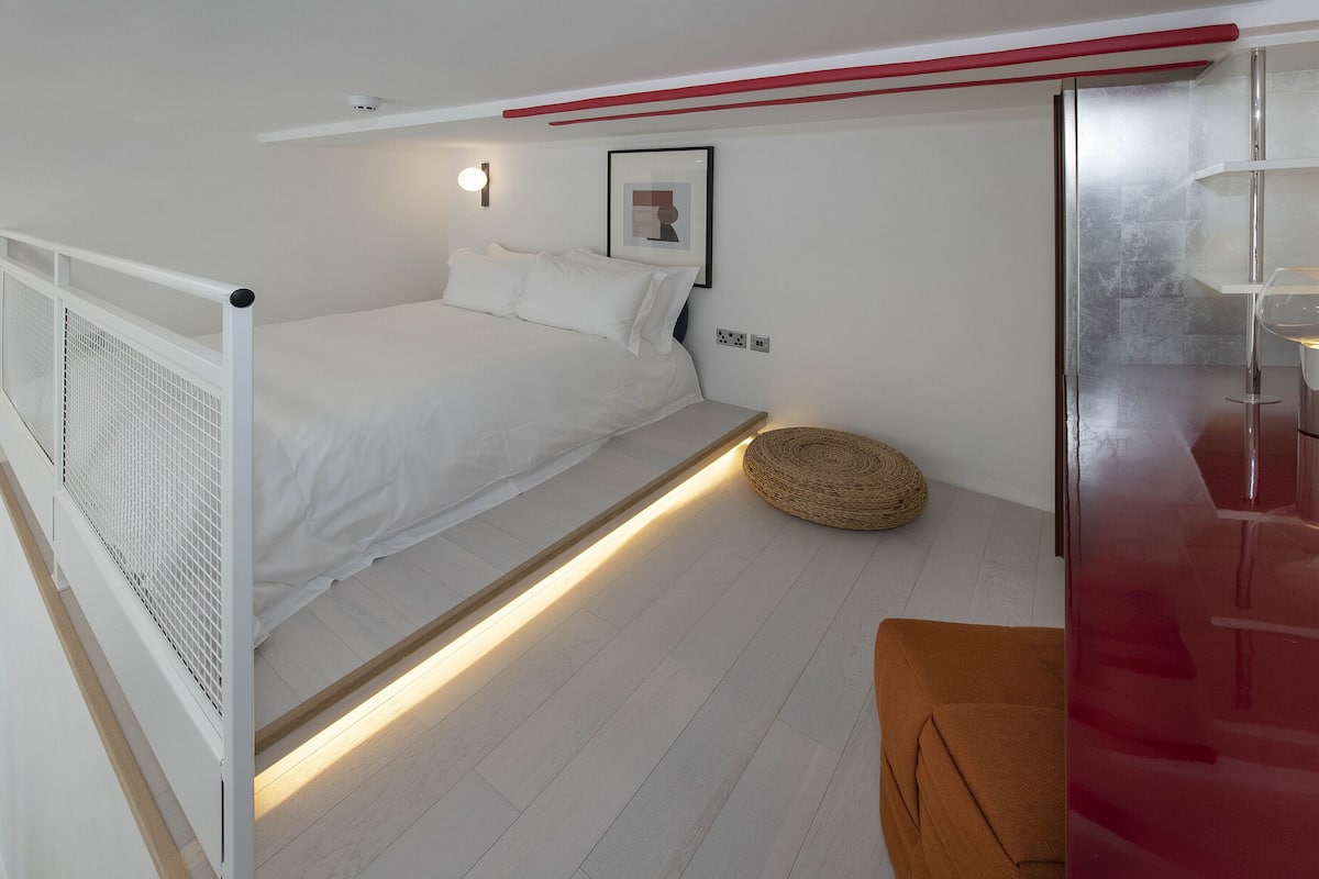 A bedroom with a bed, a bedside table with a lamp, a woven basket, and a red bench.