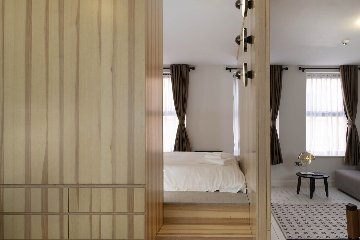 A modern hotel room with a wooden wardrobe, a bed with white linens, a small round table with a lamp, and a sofa.