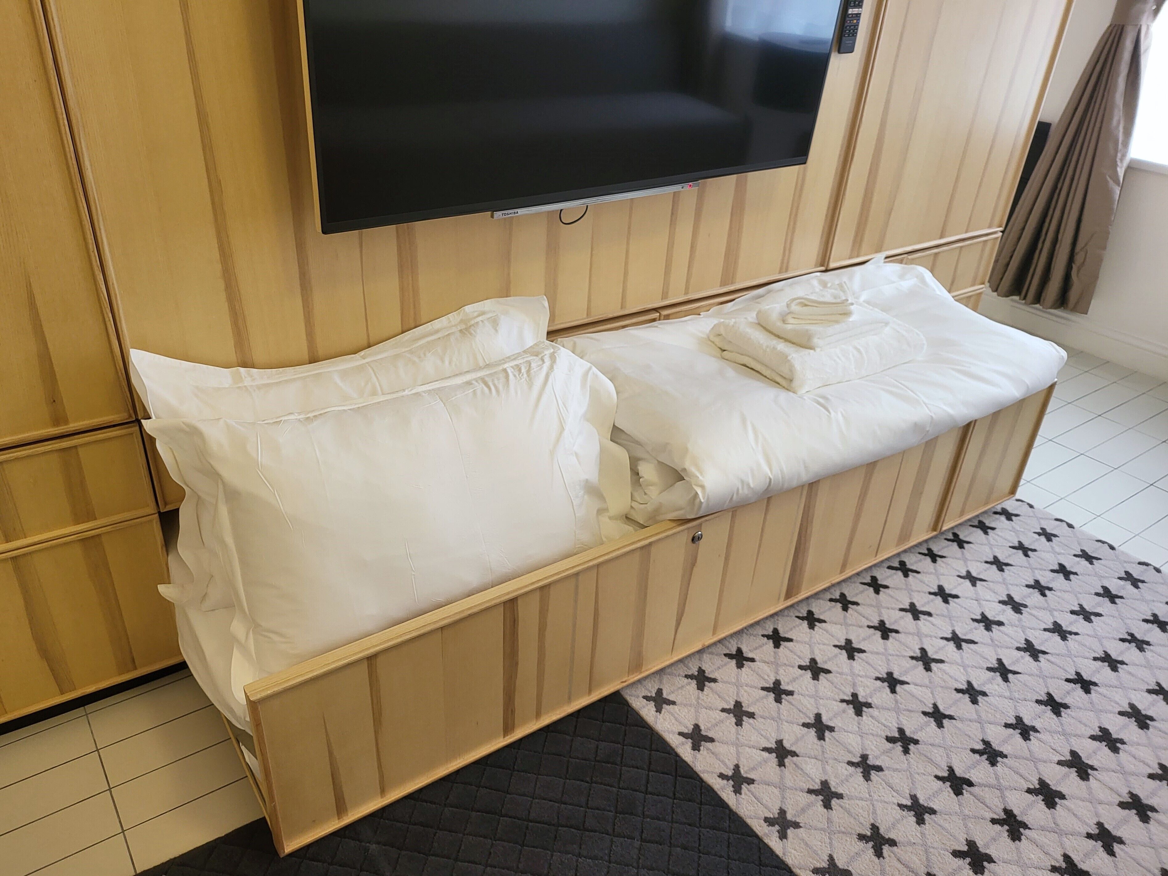 A hotel room with a bed, a flat-screen TV mounted on the wall, and a patterned rug.