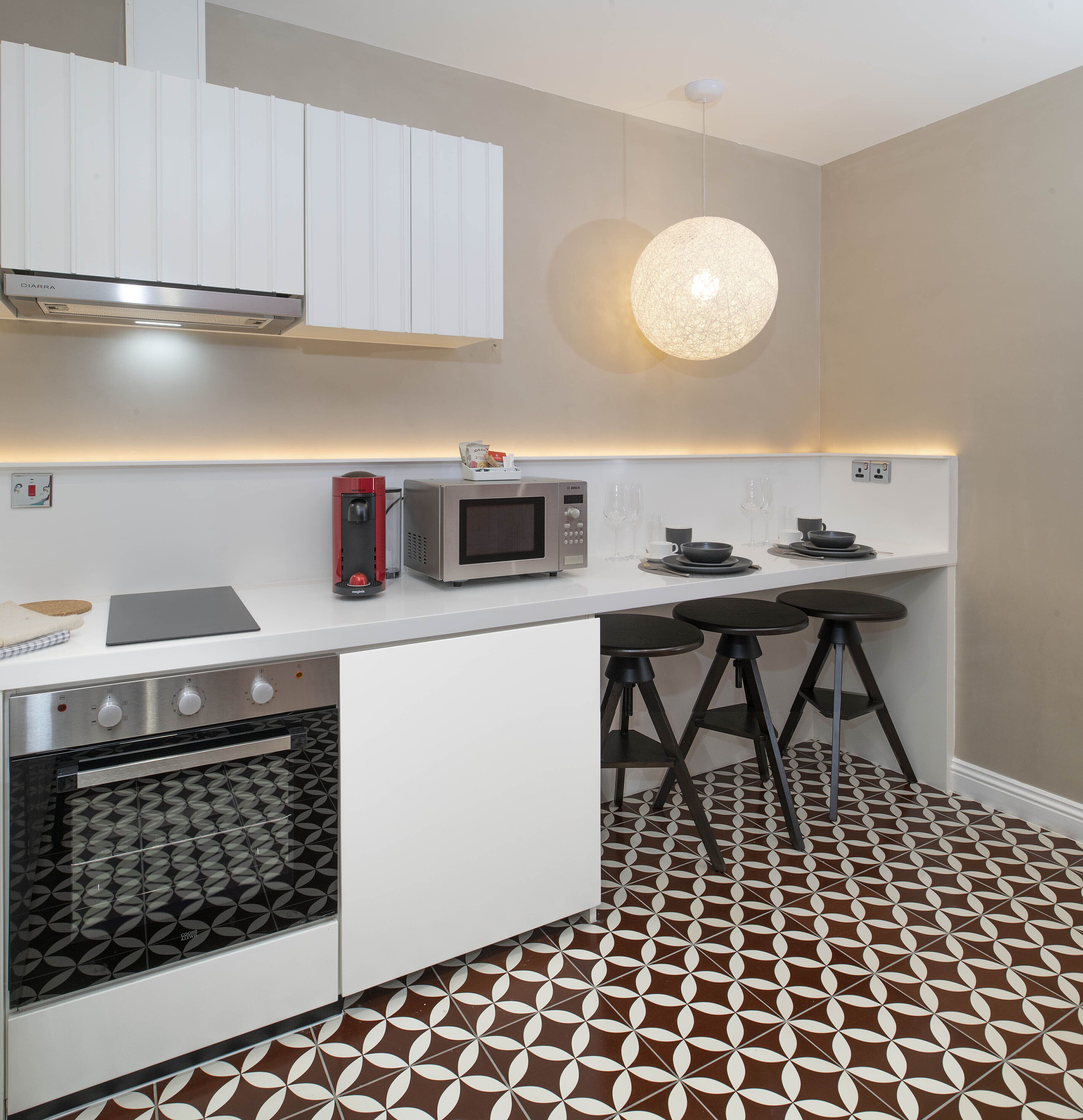A modern kitchen with white cabinetry, a built-in oven, a microwave, and a coffee machine. There are three black stools and a round pendant light above the counter.