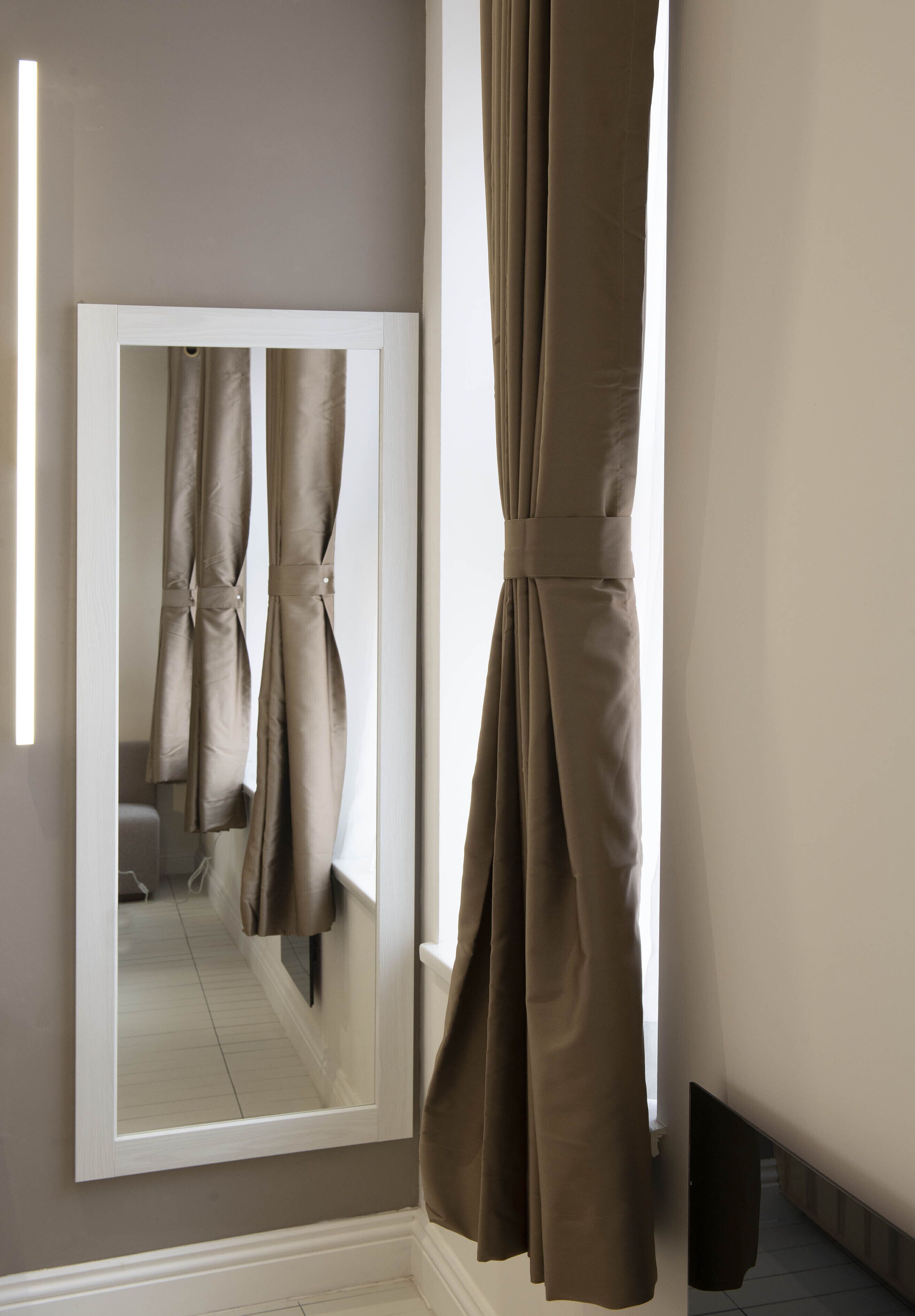 A room with a large mirror reflecting curtains, a flat-screen TV, and a white-framed mirror.