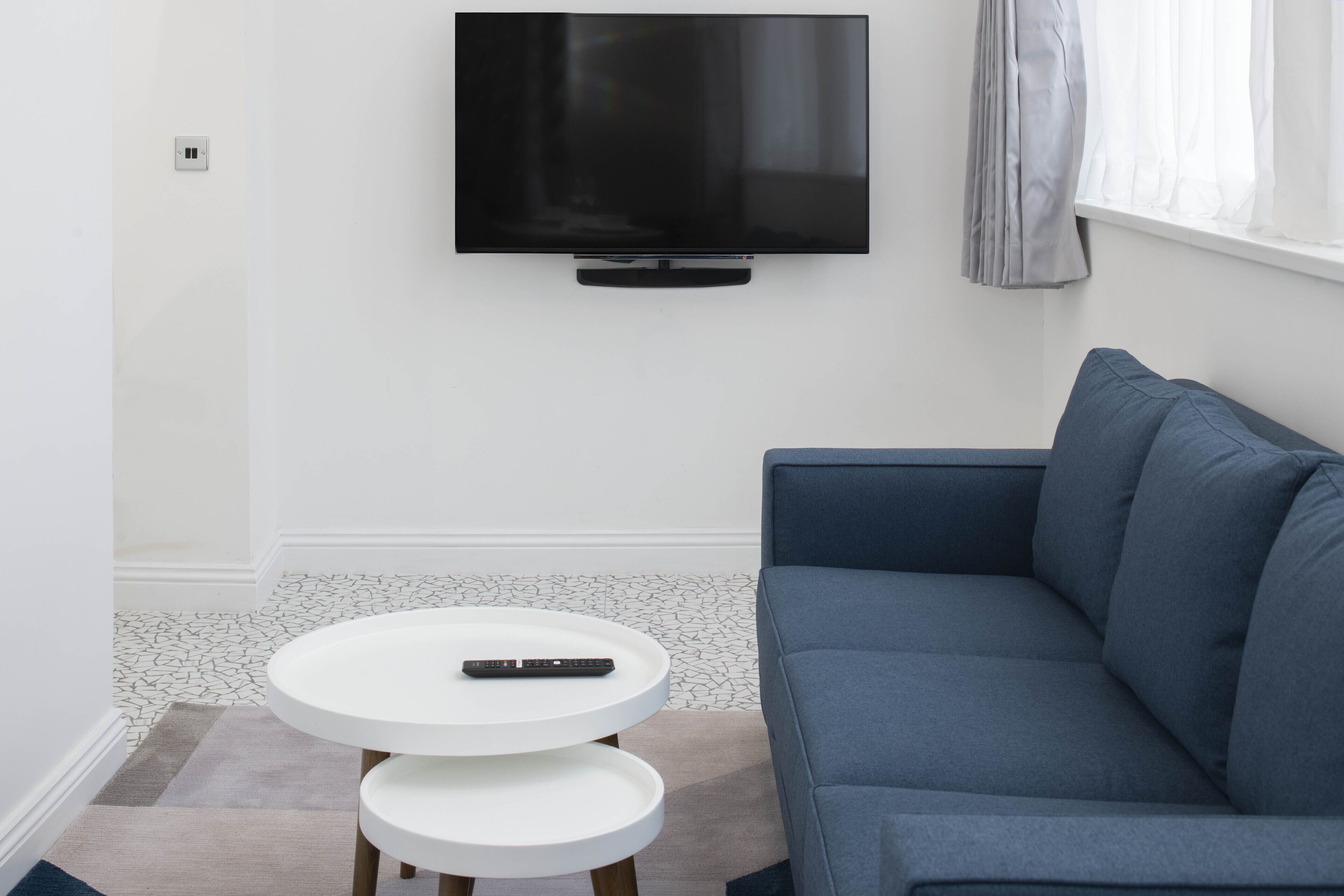 A modern living room with a flat-screen TV mounted on the wall, a blue sofa, and a white round coffee table with a remote control on it.