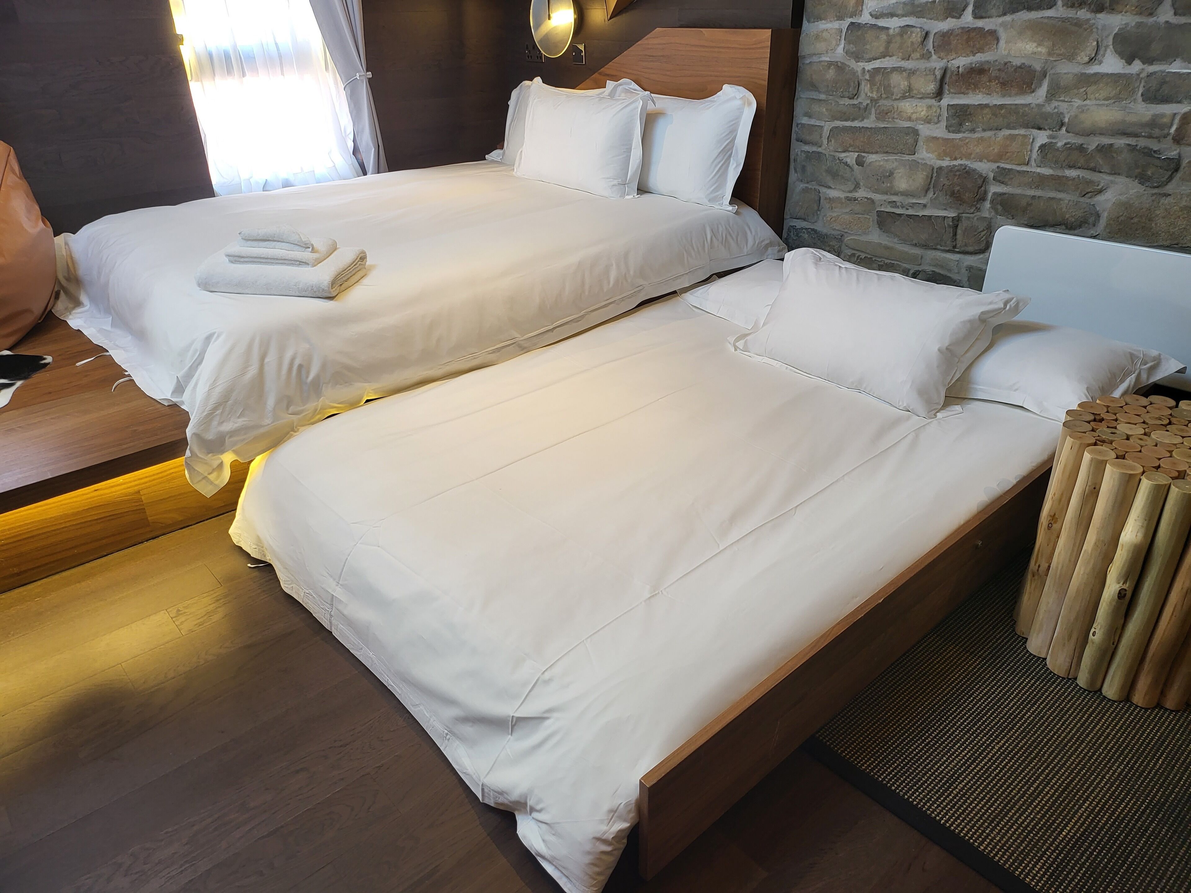 Habitación de hotel con dos camas individuales, cabeceros de madera, pared de piedra y una mesita de madera con una pila de toallas.