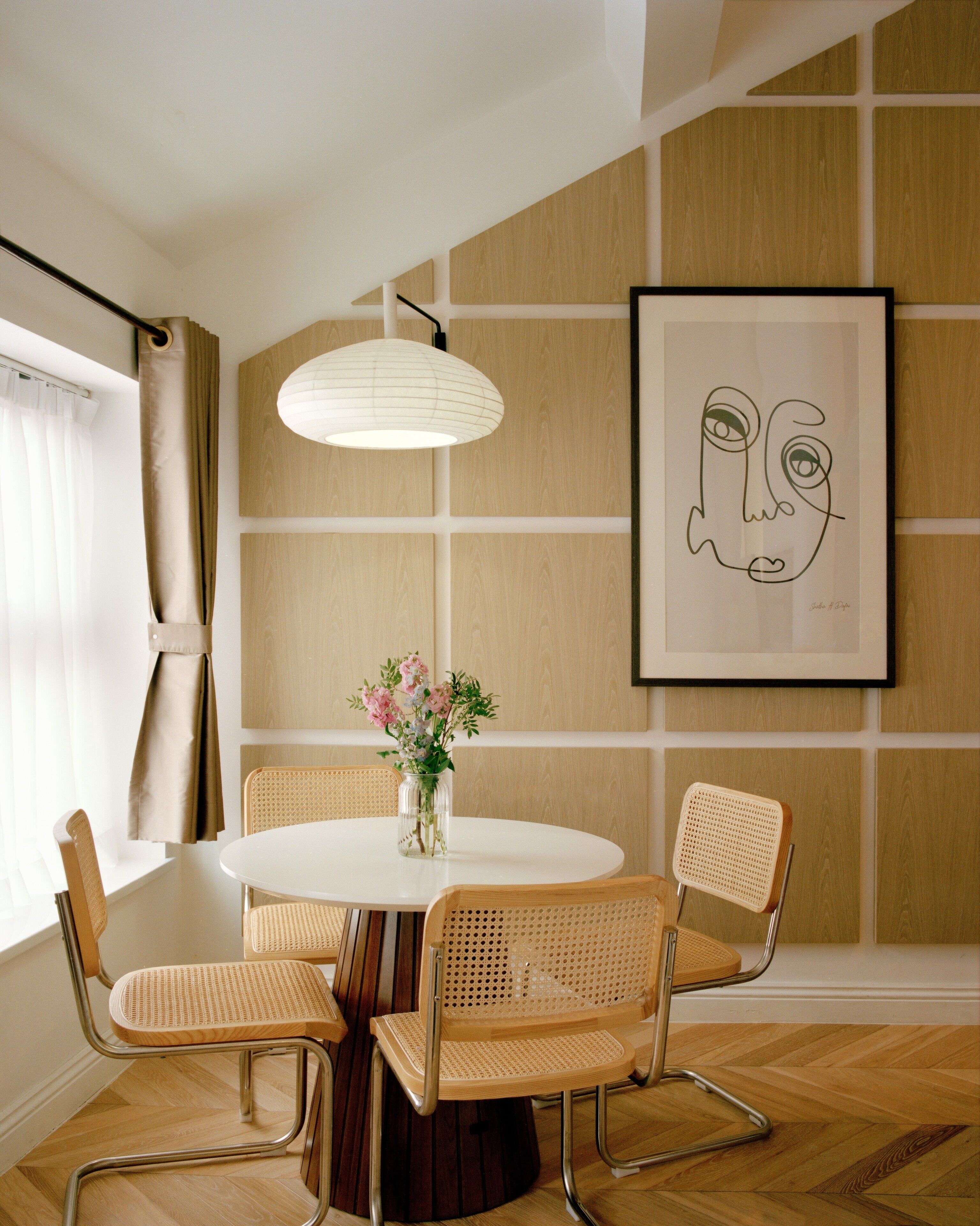Un coin repas avec une table ronde, quatre chaises en osier, un vase de fleurs, une suspension et une œuvre d’art encadrée accrochée au mur.
