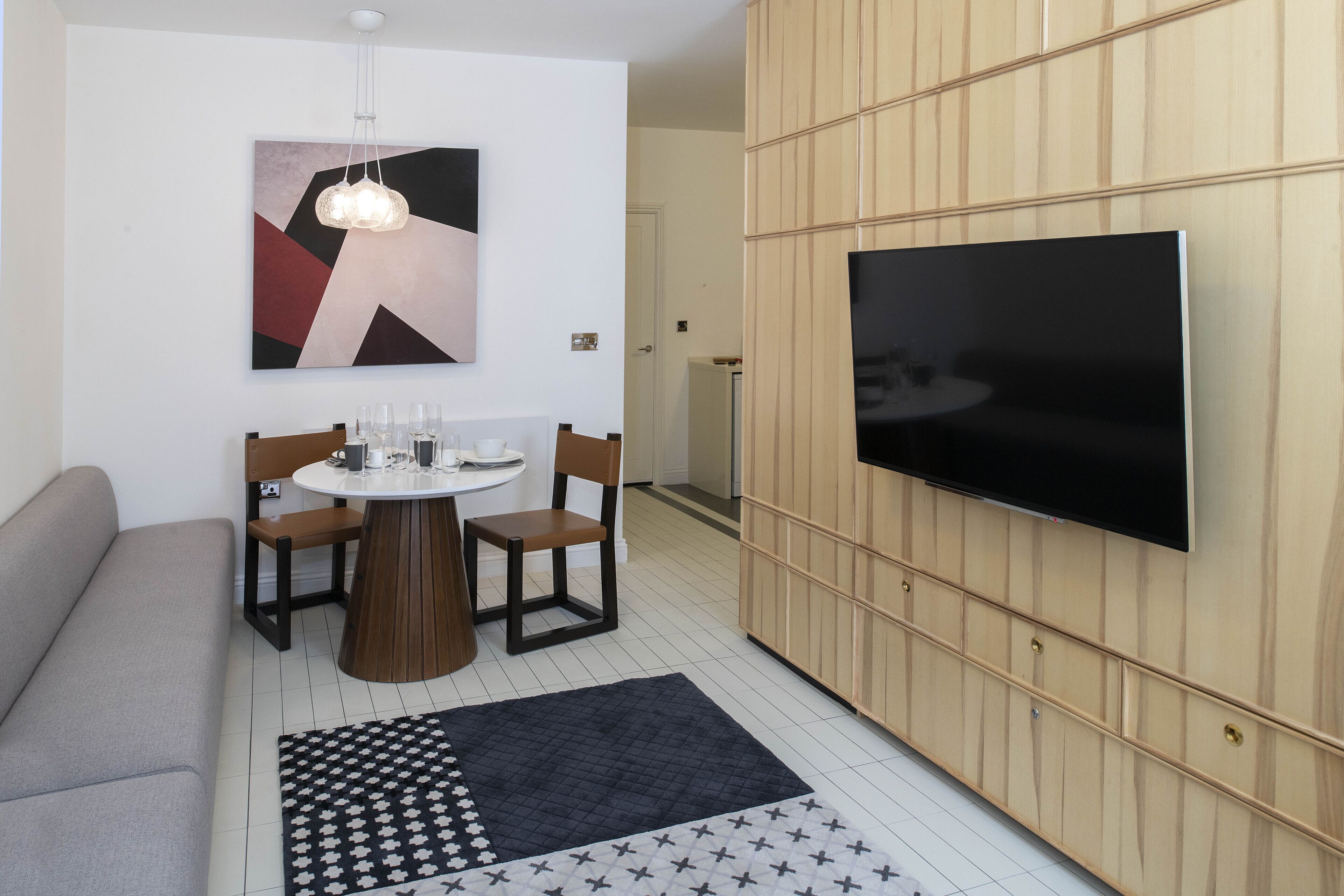 A modern living room with a sofa, a round dining table with chairs, a flat-screen TV mounted on a wooden panel wall, and a patterned rug on the floor.