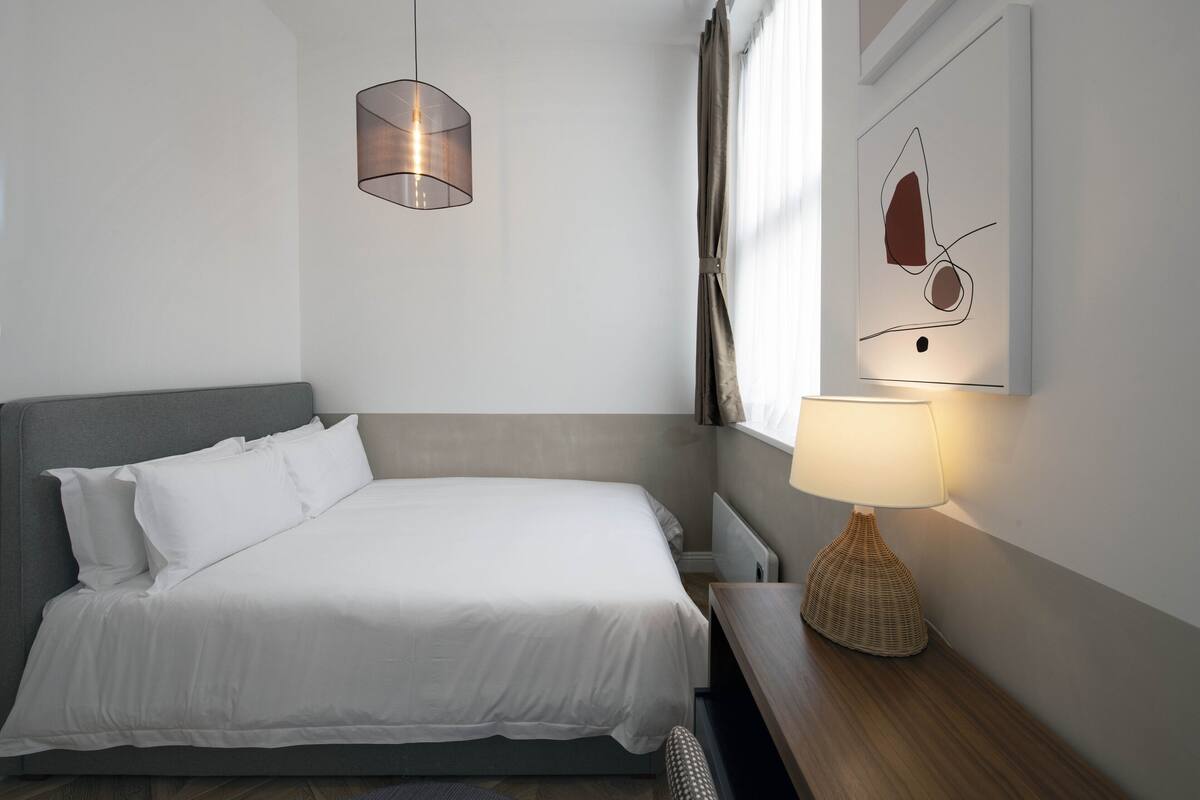 A neatly made bed with white linens, a bedside lamp, and a wall-mounted abstract art piece.