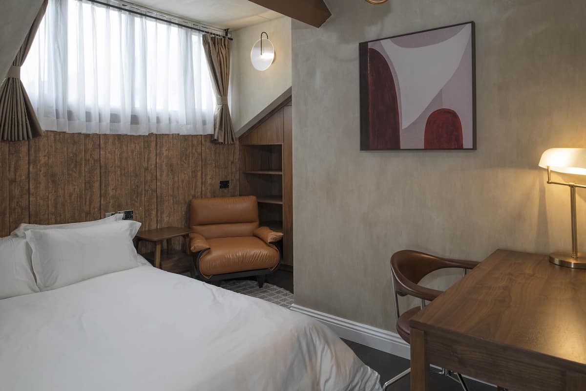 A hotel room with a bed, a desk, a chair, a wall-mounted lamp, and a painting.