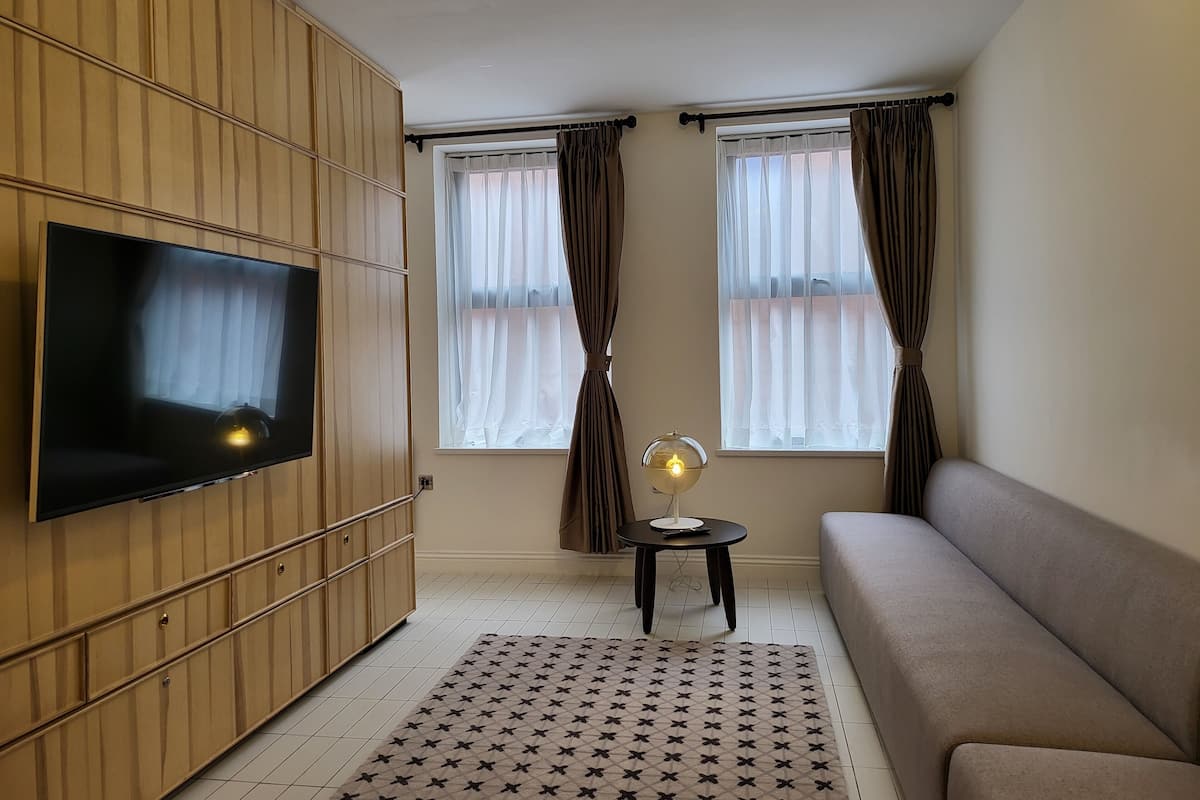 A modern living room with a flat-screen TV mounted on a wooden panel wall, a sofa, a small side table with a lamp, and two windows with sheer curtains.