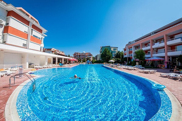 A swimming pool with a person swimming, surrounded by lounge chairs and umbrellas, with buildings in the background.