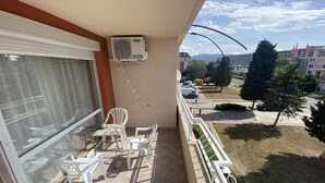 A balcony with a glass door, white plastic chairs, a table, an air conditioning unit, and a view of a street with cars and buildings. - Studio - Sleeps 4 & Access to Pools (Sunny Beach)
