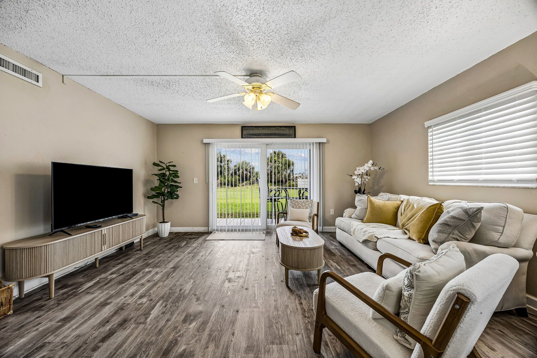 A living room with a flat-screen TV, a ceiling fan, a sofa with cushions, a coffee table, and a potted plant.