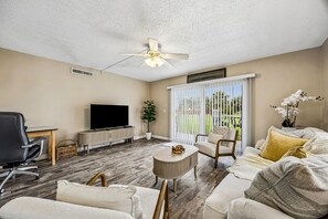 A living room with a flat-screen TV, a ceiling fan, a large window with blinds, and a plant. - SunLounger|3BR/2BA Beachfront Retreat (Saint Augustine Beach)