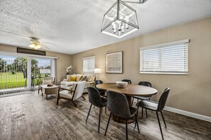A spacious living room with a dining area, large windows with blinds, a ceiling fan, and a view of a green landscape. - SunLounger|3BR/2BA Beachfront Retreat (Saint Augustine Beach)