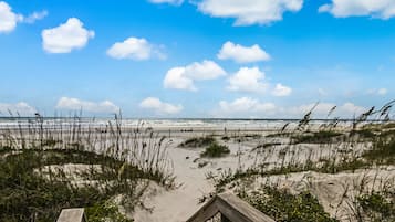 Una playa con un paseo marítimo de madera que lleva al agua, dunas de arena y un cielo despejado con nubes.