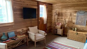 A cozy cabin interior with a wooden interior, a television mounted on the wall, a comfortable chair, a small table with books and a vase, and a colorful striped rug.