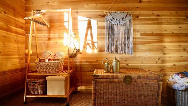 A wooden cabin interior with a wicker shelf, a macrame wall hanging, and a woven chest.