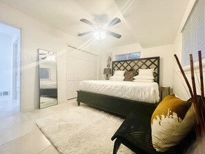 Une chambre à coucher comprenant un lit, un ventilateur de plafond, une table de chevet, une lampe et une fenêtre avec des stores.