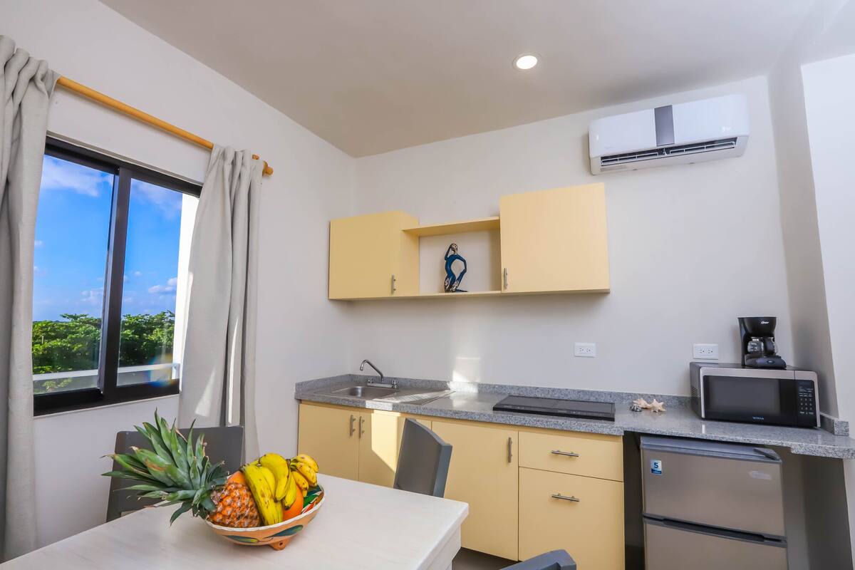A compact kitchen with a sink, microwave, and a refrigerator. There's a fruit bowl on the table and a window with curtains.