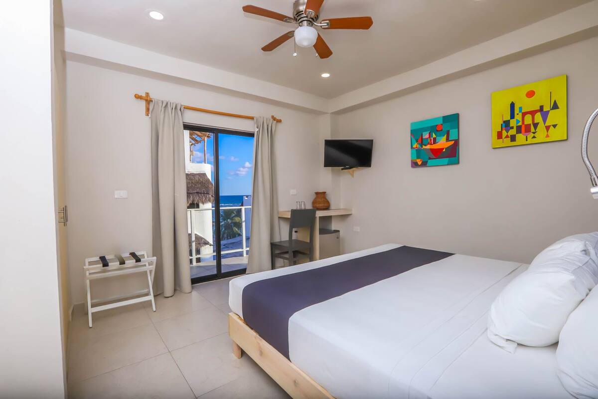 A bedroom with a bed, a desk, a chair, a TV, and a balcony with a view.