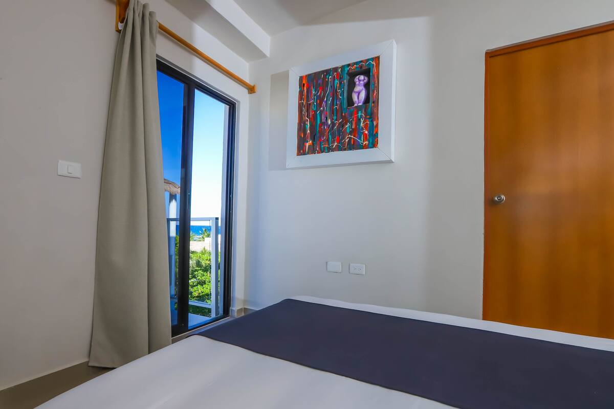 A bedroom with a bed, a window with curtains, a door, and a framed artwork on the wall.