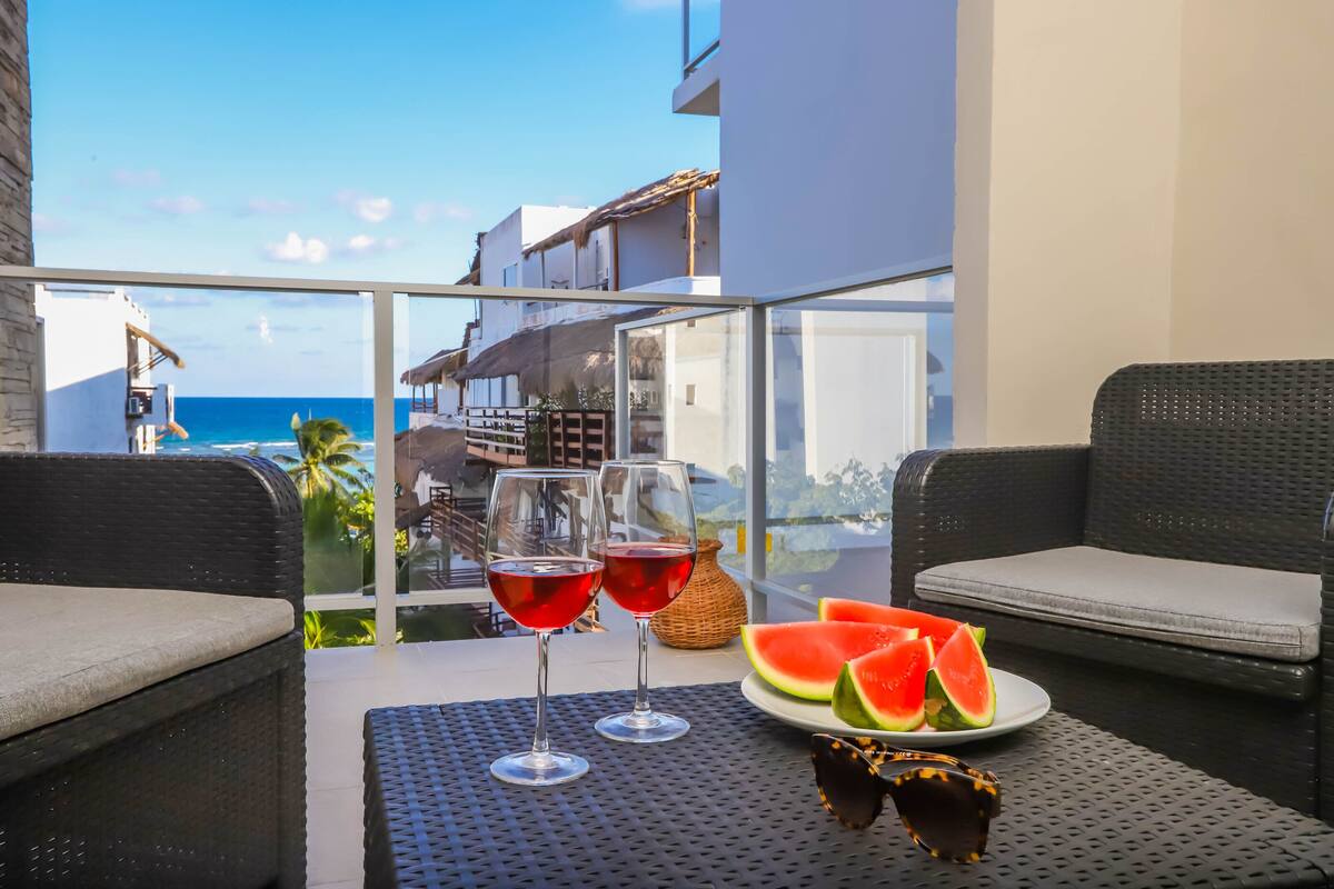 A balcony with wicker furniture, two glasses of red wine, a plate of watermelon slices, and a wicker vase.