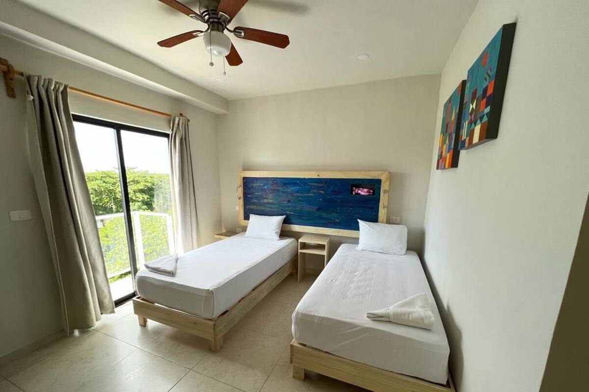 A hotel room with two single beds, a ceiling fan, a large window with curtains, and a painting on the wall.