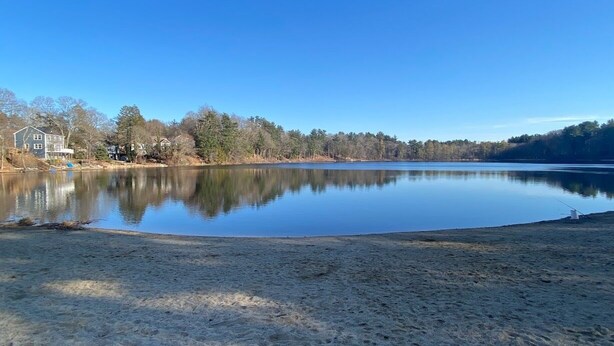A calm lake with a sandy shore, surrounded by trees and houses in the background.