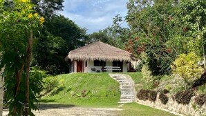 A thatched-roof house with a stone staircase leading up to it, surrounded by lush greenery and trees.