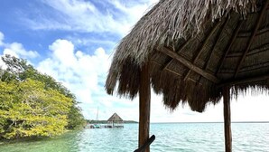 A wooden pier leading to a thatched-roof structure over turquoise water, with a clear sky and lush greenery in the background.