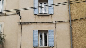Un edificio de dos pisos con fachada color beige, número 7, que presenta una puerta y contraventanas de madera, un pequeño balcón y el número 5 visible a la izquierda.