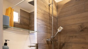 Un baño moderno con pared de madera, estante de vidrio, lavamanos blanco y ducha con cabezal de ducha manual.