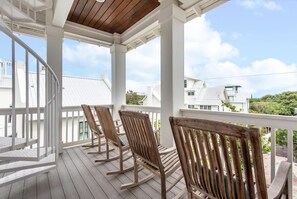 A wooden deck with white railings and rocking chairs, overlooking a residential area with white houses.
