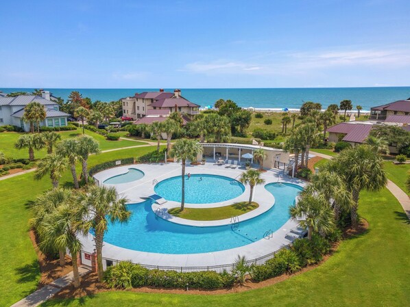 A resort with a large central pool, multiple buildings, and a view of the ocean.