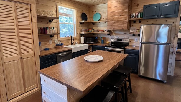 A modern kitchen with wooden cabinetry, a central island with bar stools, stainless steel appliances, and built-in shelves. - Beautiful rustic cabin on the shores of Greer's Ferry Lake (Higden)