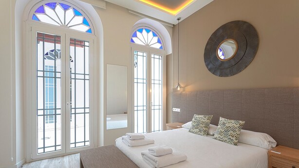 A modern hotel room with a large bed, two nightstands, a round mirror, and a view through the arched windows. - Fresh Velazquez Apartment E (Málaga)