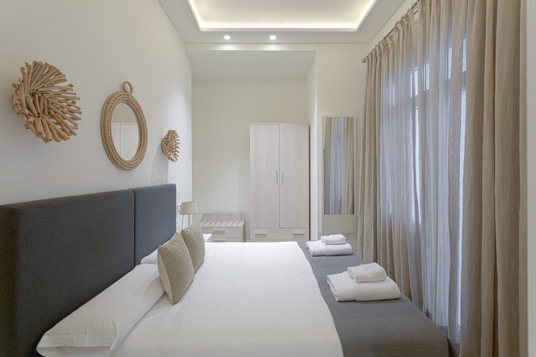 A modern hotel room with a large bed, a wardrobe, a mirror, and a view of the outdoors through sheer curtains.