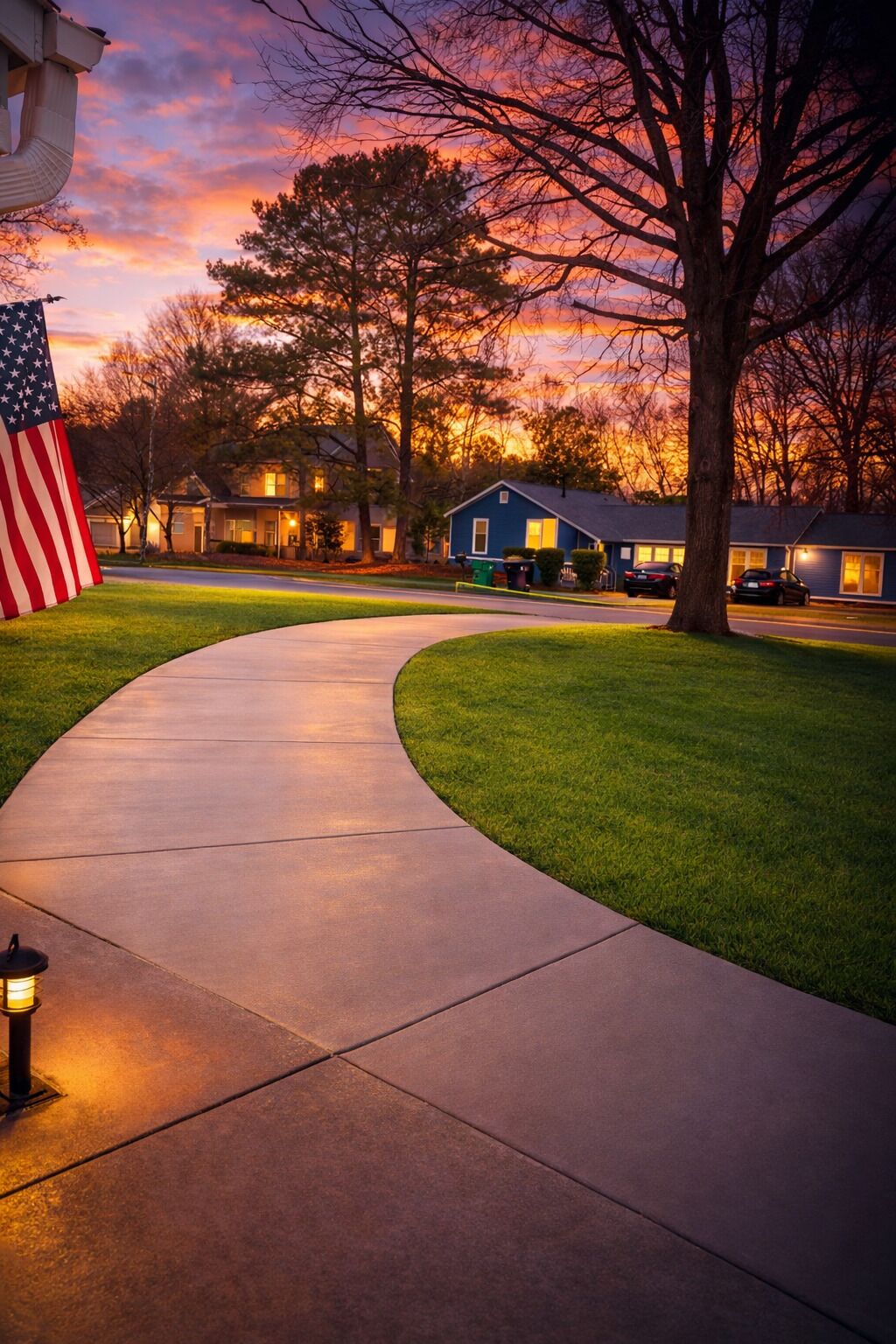 A suburban neighborhood at sunset with a curved sidewalk, a flag, and houses with lit windows.