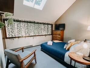 Un dormitorio con cama, televisión, cómoda, silla y ventana con plantas colgantes.