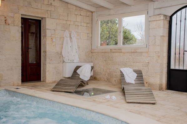 A stone-walled indoor pool area with two wooden loungers, a glass-enclosed hot tub, and a window overlooking greenery. - Trullo Panoramico - Micele Holiday Houses (Locorotondo)