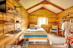 A cozy log cabin room with a bed, a chair, a robe rack, and a shelf with a WiFi sign.